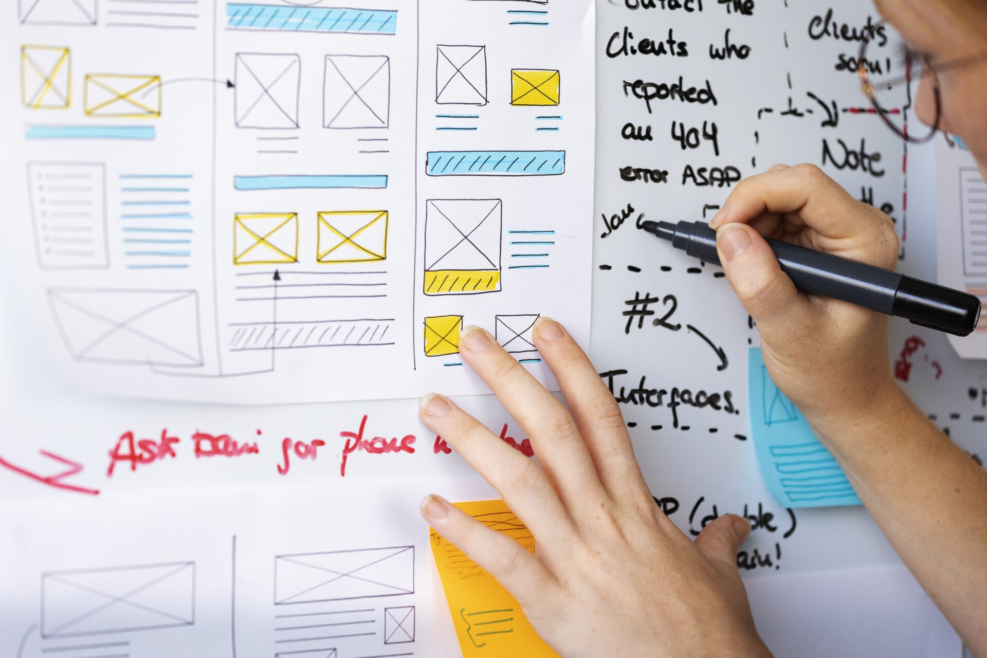 Female web designer is making notes with UX design mockups pinned to a wall.